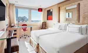 Hotellihuone, jossa on kaksi yhden hengen vuodetta, kirjoituspöytä ja suuri ikkuna, josta on näkymä kaupunkiin. (Tekoälyn luoma teksti)