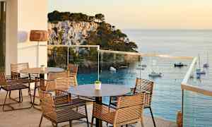 Terassi, jossa pöytiä ja tuoleja, näkymä merelle ja veneitä kalliorannikolla. (Tekoälyn luoma teksti)