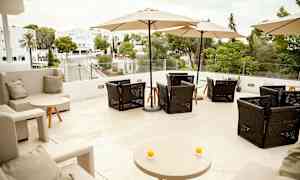 Terassi, jossa istuimia ja aurinkovarjoja, näkymä vihreisiin alueisiin ja rakennuksiin. (Tekoälyn luoma teksti)