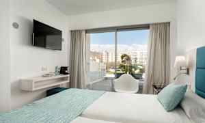 Hotellihuone, jossa kaksi vuodetta vaaleilla lakanoilla ja turkooseilla päiväpeitteillä. Tuoli on lasioven edessä, joka johtaa parvekkeelle. Seinään kiinnitetty televisio on hyllyn vieressä, jossa on vesipulloja ja kahvinkeitin. (Tekoälyn luoma teksti)