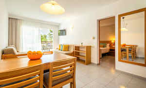 Valoisa olohuone, jossa ruokapöytä, tuolit, sohva ja parveke näkymällä vehreään. Liittyvä makuuhuone näkyvissä. (Tekoälyn luoma teksti)