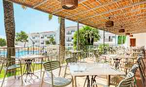 Terassi, jossa pöydät ja tuolit, uima-allas ja hotellirakennukset taustalla. (Tekoälyn luoma teksti)