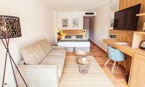 Hotellihuone, jossa on sohva, parivuode, kirjoituspöytä tuolilla ja televisio. (Tekoälyn luoma teksti)