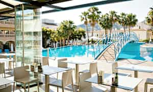 Ravintola-alue ulkoistuimineen altaan äärellä, palmuja ja rakennuksia ympärillä. Sininen jalankulkusilta ylittää altaan. (Tekoälyn luoma teksti)