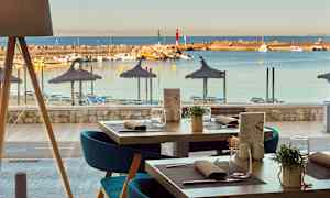 Näkymä ravintolasta rannalle, jolla on aurinkovarjoja ja aurinkotuoleja, sekä pienvenesatamaan veneineen. (Tekoälyn luoma teksti)