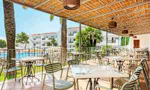 Ulkoterassi, jossa on pöytiä ja tuoleja katoksen alla, näkymä uima-altaalle ja hotellirakennuksiin. (Tekoälyn luoma teksti)