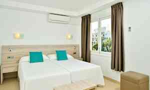 Hotellihuone, jossa on kaksi vierekkäistä yhden hengen vuodetta, valkoinen sisustus ja turkoosit tyynyt, sekä ikkuna näkymällä. (Tekoälyn luoma teksti)