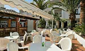 Ulkoterassi, jolla on valkoisia tuoleja ja pöytiä markiisin alla, palmujen ja kasvillisuuden ympäröimänä. (Tekoälyn luoma teksti)