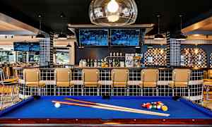 Biljardipöytä sinisellä veralla baarin edessä, jossa on tuoleja ja televisioruutuja. (Tekoälyn luoma teksti)