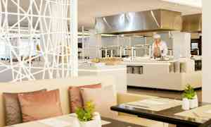 Moderni ravintolatila, jossa on valkoisia huonekaluja, pastellisävyisiä tyynyjä ja pöydillä kasveja, taustalla näkyy avoin keittiö. (Tekoälyn luoma teksti)