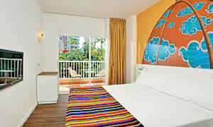 Hotellihuone, jossa on parivuode ja värikäs päiväpeite, kuvioitu seinämaalaus sängyn takana, ikkuna, josta on näkymä parvekkeelle ja vehreyteen. (Tekoälyn luoma teksti)