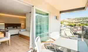 Hotellihuone, jossa on kaksi vuodetta ja istuinhuonekalu, siihen liittyvä parveke, jossa on pöytä ja tuolit sekä näkymä vehreälle kukkulalle. (Tekoälyn luoma teksti)