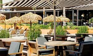 Ulkotila, jossa pöytiä, tuoleja ja aurinkotuoleja olkikatosten alla. (Tekoälyn luoma teksti)