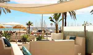 Näkymä rauhallisesta istuinalueesta, jossa on valkoisia huonekaluja ja sinisiä tyynyjä, trooppisten kasvien ympäröimänä, taustalla ranta ja venesatama. (Tekoälyn luoma teksti)