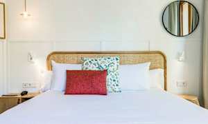 Hotellihuone, jossa on pedattu parivuode koristeltu valkoisilla tyynyillä ja punaisella koristeellisella tyynyllä kukkakuosin edessä. Sängynpääty on punottua puuta. Pyöreä peili ja seinävalaisimet näkyvät vaalealla taustalla. (Tekoälyn luoma teksti)