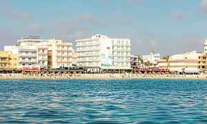 Näkymä merestä rannalle, jossa on useita rakennuksia ja ihmisiä auringossa. (Tekoälyn luoma teksti)