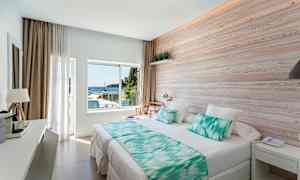 Valoisa hotellihuone, jossa suuri sänky, seinässä puupaneeli ja ikkuna, josta näkyy meri. (Tekoälyn luoma teksti)