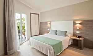 Hotellihuone, jossa on parisänky, kaksi tyynyä ja vihreä peitto. Sänky sijaitsee lähellä parveketta, jossa on lasiovet. (Tekoälyn luoma teksti)