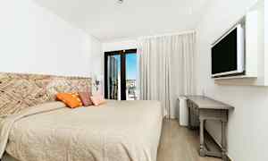 Hotellihuone, jossa on suuri sänky, koristeellisia tyynyjä, seinään kiinnitetty televisio ja kirjoituspöytä. Parvekkeen ovet avaavat näkymän. (Tekoälyn luoma teksti)