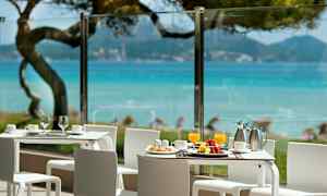 Kuvassa on terassi, jossa on valkoiset pöydät ja tuolit sekä näkymä siniselle merelle ja vehreään ympäristöön. (Tekoälyn luoma teksti)