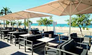 Ulkokahvila, jossa on pöytiä, tuoleja ja aurinkovarjoja, näkymä hiekkarannalle ja merelle. (Tekoälyn luoma teksti)