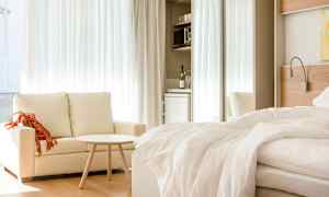 Vaalea hotellihuone, jossa on sohva, pieni pöytä ja pedattu sänky. Taustalla on minikeittiö jääkaapilla. (Tekoälyn luoma teksti)