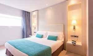 Vaalea hotellihuone, jossa on parivuode, kaksi turkoosia tyynyä, pehmeä huopa ja seinälamput sängyn molemmilla puolilla. (Tekoälyn luoma teksti)