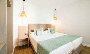 Hotellihuone, jossa kaksi pedattua yhden hengen vuodetta, vihreät vuodetekstiilit ja ripustetut valaisimet. (Tekoälyn luoma teksti)