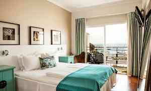 Hotellihuone, jossa on parivuode ja valkoiset ja siniset vuodevaatteet, seinätaidetta sängyn yläpuolella ja parveke merinäköalalla. (Tekoälyn luoma teksti)