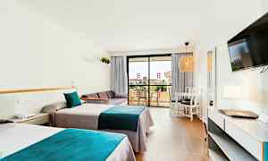 Hotellihuone, jossa on kaksi yksittäistä vuodetta sinisillä ja harmailla vuodevaatteilla, sohva, pöytä tuoleineen sekä parveke taustalla. (Tekoälyn luoma teksti)