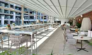 Moderni ja valoisa terassi- tai ravintolatilaksi sisustettu alue, jossa on lasiseinät, rivi pöytiä ja tuoleja sekä koristekasveja. (Tekoälyn luoma teksti)