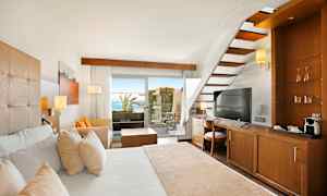 Hotellihuone puukalusteilla, valkoinen sänky, portaat ja näkymä terassille, joka avautuu merelle. (Tekoälyn luoma teksti)