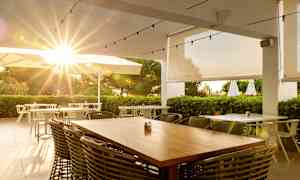 Ulkoterassiravintola, auringonsäteitä päivänvarjojen ja vehreyden läpi. (Tekoälyn luoma teksti)