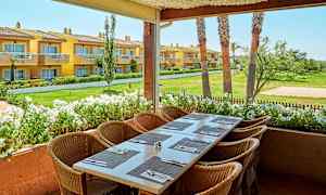 Kuva ulkoterassista, jossa on katettu pöytä ja tuolit, näkymä puutarhaan ja keltaisiin rakennuksiin taustalla. (Tekoälyn luoma teksti)