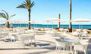 Ulkoilualue, jossa on valkoisia kalusteita ja aurinkovarjoja, sekä palmupuita taustalla meren äärellä. (Tekoälyn luoma teksti)