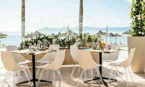 Kuva ulkoilmaravintolasta, jossa on valkoiset tuolit ja pöydät, merinäköalalla ja taustalla aurinkovarjoja. (Tekoälyn luoma teksti)