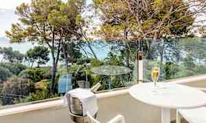 Parveke, jolta on näkymä puihin ja merelle. Parvekkeella on valkoinen pöytä, jonka päällä on samppanjalasi, sekä teline, jossa on jäähdytysastia samppanjapullolle. (Tekoälyn luoma teksti)