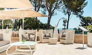 Ulkona oleskelualue, jossa valkoiset punotut kalusteet, beige tyynyt ja aurinkovarjoja. Taustalla vihreitä puita ja merinäköala. (Tekoälyn luoma teksti)