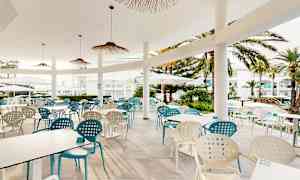 Ulkoterassi pöytineen ja tuoleineen, jota ympäröivät palmut ja valkoiset rakennukset. (Tekoälyn luoma teksti)