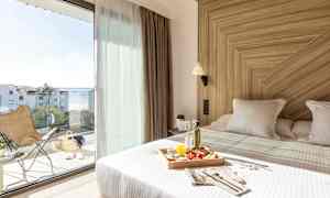 Valoisa hotellihuone, jossa on pedattu sänky ja aamiaistarjotin. Suuret lasiovet avautuvat parvekkeelle, jolta on näkymä merelle. (Tekoälyn luoma teksti)