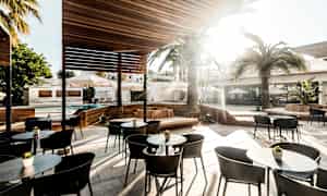 Ulkoterassi, jossa on istuimia, pöytiä ja aurinkosuojia puisen katon alla. Taustalla näkyy palmuja ja aurinkovarjoja. (Tekoälyn luoma teksti)