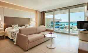 Hotellihuone, jossa on sänky, sohva, pöytä ja näkymä merelle lasiovien kautta. (Tekoälyn luoma teksti)