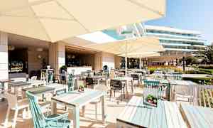 Ulkoterassiravintola, jossa on pöytiä ja päivänvarjoja, taustalla näkyy rakennuksia. (Tekoälyn luoma teksti)