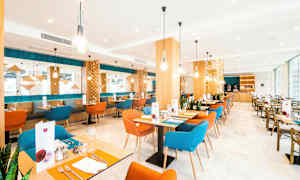 Kuva nykyaikaisesta ravintolasta, jossa on värikkäitä tuoleja, puupöytiä ja koristeellisia valaisimia. (Tekoälyn luoma teksti)