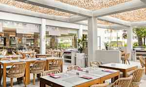 Valoisa ravintola, jossa on puupöydät ja rottinkituolit, sijaitsee modernissa sisätilassa koristeellisen katon ja ulkopuolelle avautuvien lasiseinien kanssa. (Tekoälyn luoma teksti)