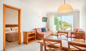 Hotellihuoneisto, jossa on pöytä, tuoleja, sohva ja makuuhuone näkyvissä avoimen oven läpi. (Tekoälyn luoma teksti)