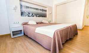 Hotellihuone, jossa on suuri sänky, ruskea päiväpeite ja valkoiset lakanat, puulattia ja seinäkoriste mustavalkoisen kuvan muodossa. (Tekoälyn luoma teksti)