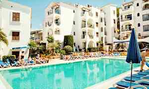 Uima-allasalue, jossa on aurinkotuoleja kirkkaansinisen altaan ympärillä, valkoisen rakennuksen edessä. (Tekoälyn luoma teksti)