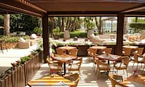 Avoin ravintola, jossa on pöytiä ja tuoleja, vehreyden ympäröimänä ja näkymä puutarhaan ja uima-altaalle. (Tekoälyn luoma teksti)