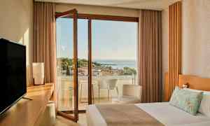 Hotellihuone, jossa on sänky, työpöytä, televisio ja avoin parvekkeen ovi, joka johtaa parvekkeelle merinäköalalla ja puiden ympäröimänä. (Tekoälyn luoma teksti)